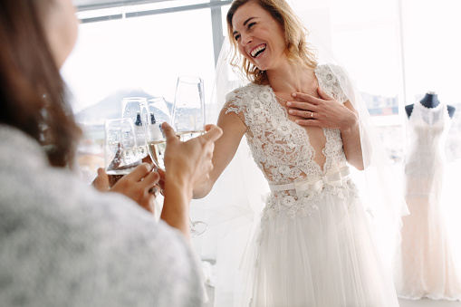 Cheerful young woman in wedding gown toasting champagne with friends in bridal Boutique. Beautiful bride in elegant wedding dress clinking glasses of champagne with her friends and smiling in wedding fashion shop.