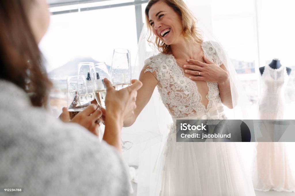 Bride toasting champagne with friends in bridal boutique Cheerful young woman in wedding gown toasting champagne with friends in bridal Boutique. Beautiful bride in elegant wedding dress clinking glasses of champagne with her friends and smiling in wedding fashion shop. Wedding Dress Stock Photo