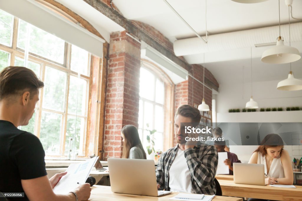 Diverse people focused on work in modern loft co-working space Diverse people focused on work in modern loft co-working space interior, businessman talking on phone while other businesspeople using laptops working with documents sitting at shared office desks Office Stock Photo