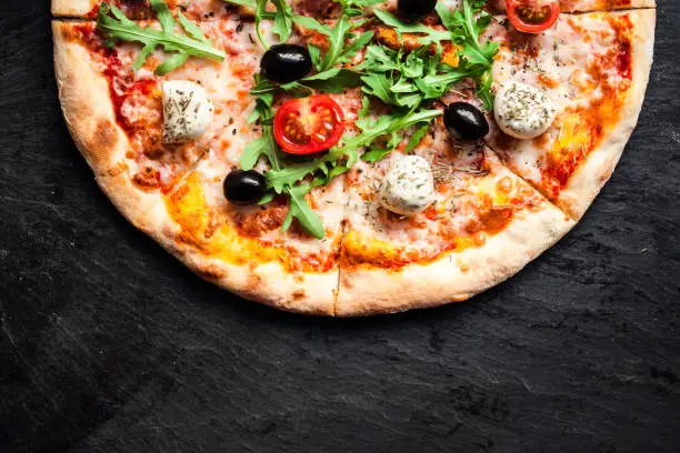 Photo of Pizza with tomatoes, mozzarella cheese and arugula. Pizza menu.Top view with copy space on dark stone table