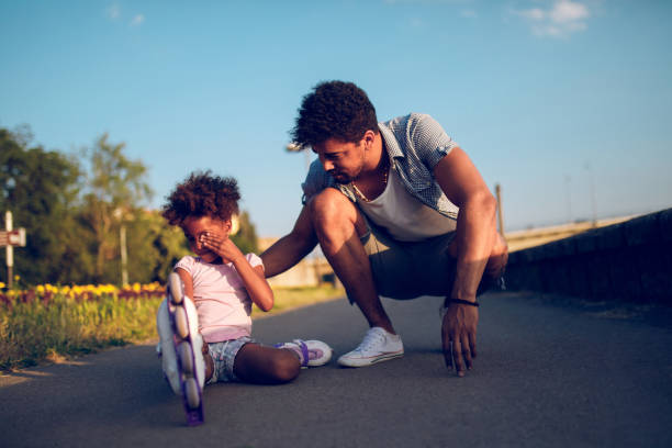 vater tröstet seine tochter, die beim rollschuhlaufen fiel - family african ethnicity black african descent stock-fotos und bilder