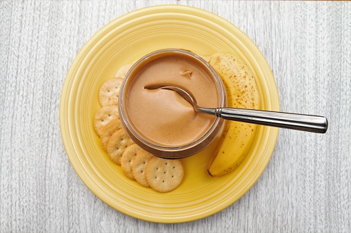 Peanut butter in open jar from above on countertop wit banana and crackers on plate below. spoon in peanut butter.