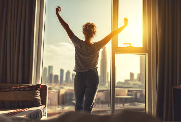 feliz mujer se extiende y abre cortinas en ventana de mañana - levantarse fotografías e imágenes de stock