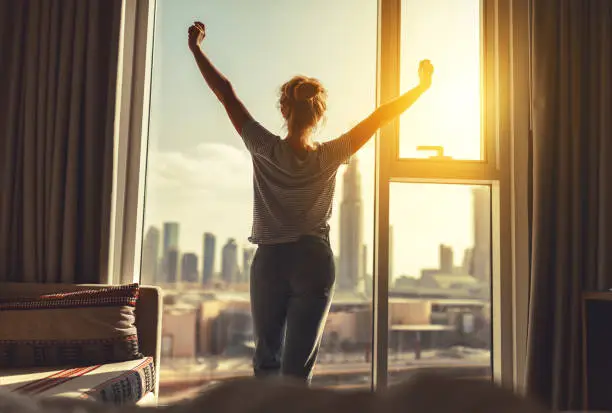 happy woman stretches and opens the curtains at window in morning