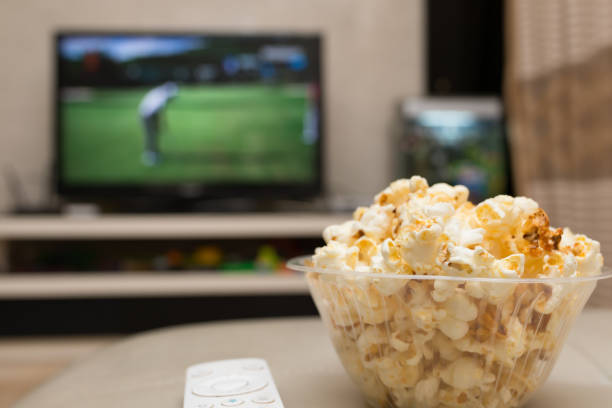 popcorn and remote control on sofa with a TV broadcasting golf match on background stock photo