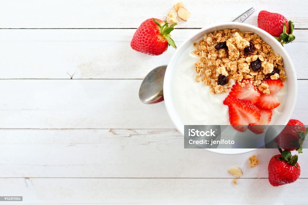 Yogurt with strawberries & granola, flat lay, side orientation over white wood. Bowl of yogurt with strawberries and granola, over a white wood background. Flat lay, side orientation. Yogurt Stock Photo