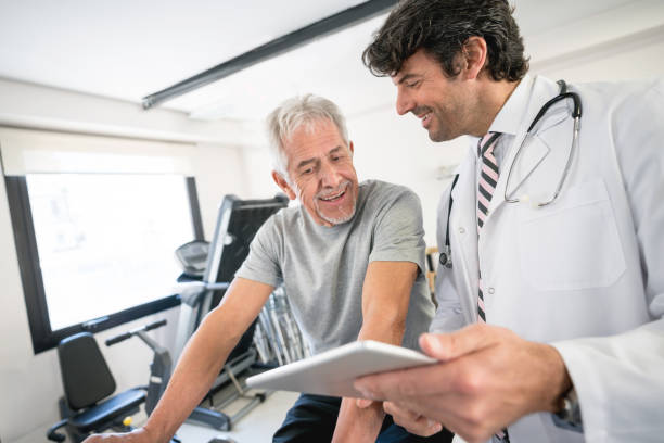 Senior patient asking questions to his physician about his recovery plan Senior patient asking questions to his physician about his recovery plan while doing therapy on a static bicycle both looking happy and smiling exercise machine photos stock pictures, royalty-free photos & images