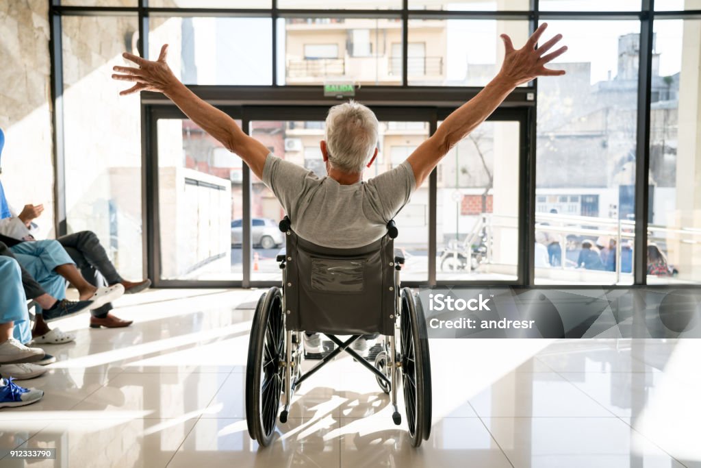 Unrecognizable senior patient leaving the clinic on a wheelchair with his arms up Backview of unrecognizable senior patient leaving the clinic on a wheelchair with his arms up Hospital Stock Photo