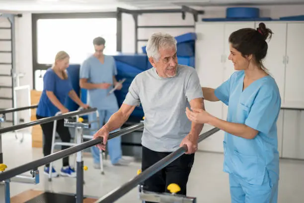 Cheerful senior man using parallel bars to walk and physiotherapist smiling at him very happy while another patient and professional are walking at the background