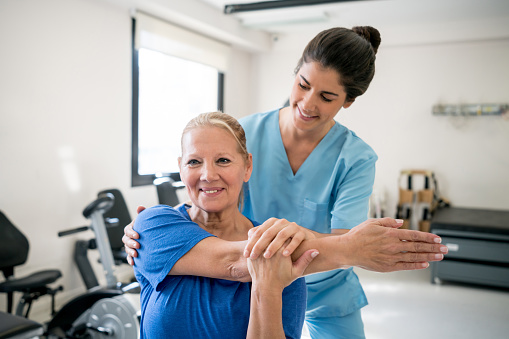 Friendly physical therapist holding senior patient while she stretches her shoulder both looking happy and smiling