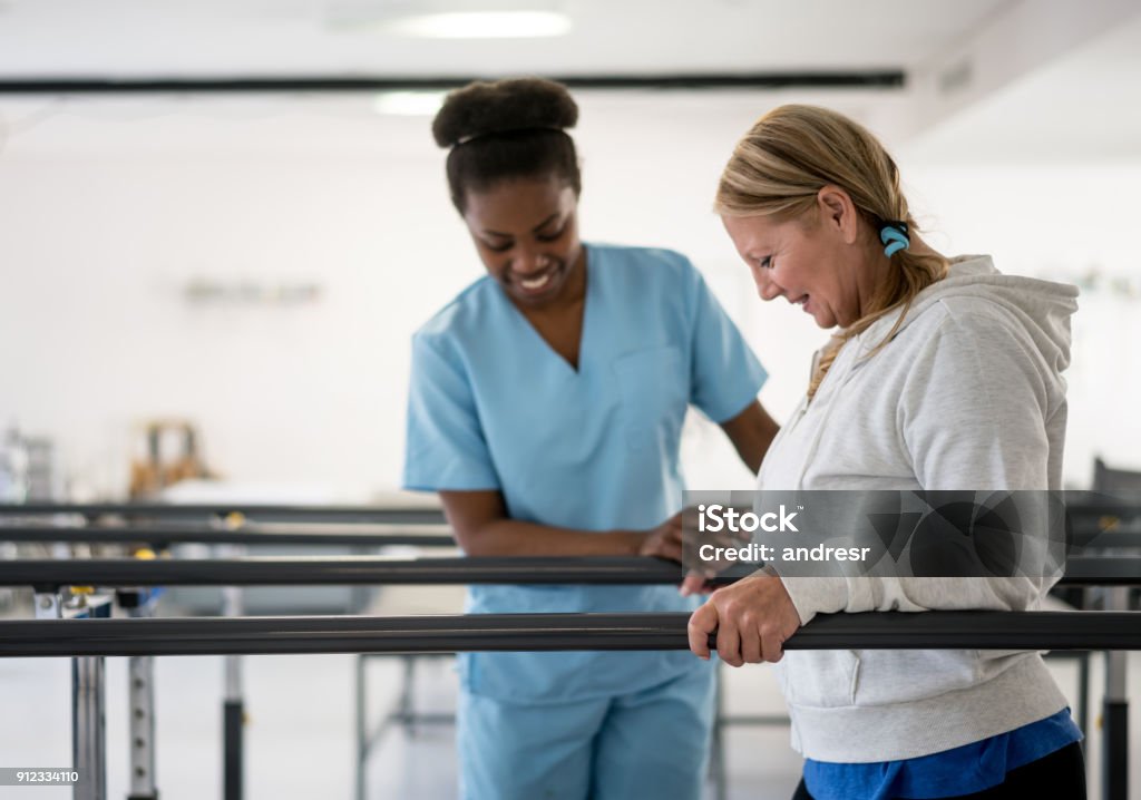Lycklig senior kvinna promenader mellan Barr på physiotheray med hennes terapeut bredvid henne - Royaltyfri Fysioterapi Bildbanksbilder