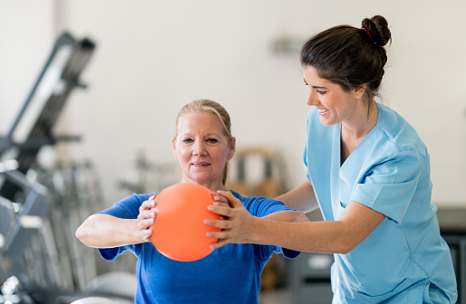 Senior woman at physical rehabilitation doing a chest workout with a ball and therapist helping her both smiling