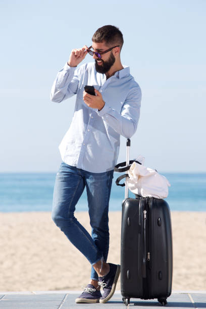 istock Full length man standing on beach with suitcase and mobile phone 912334196