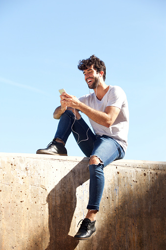 Full body portrait of cheerful man sitting on wall with headphones and cellphone