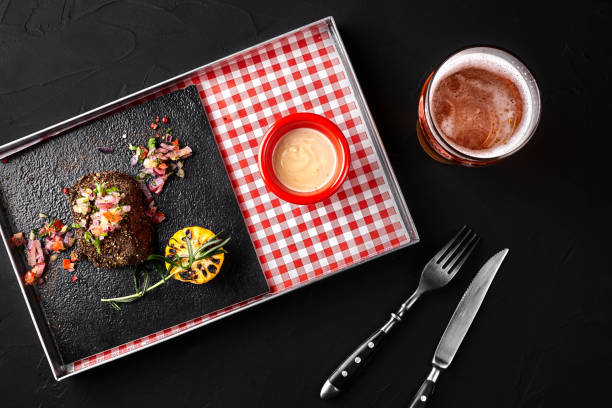 Medium rare grilled Beef steak Ribeye with corn, rosemary, onion and white sauce on a metal tray on a black background