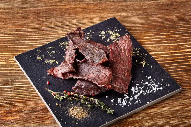 Dried peppered beef jerky cut in strips on the black board on wooden background. Still life. Copy space