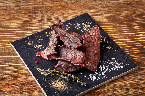 secos salpicado carne seca cortada em tiras no quadro negro sobre fundo de madeira - beef jerky meat smoked - fotografias e filmes do acervo