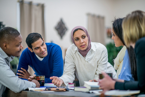 A diverse group of entrepreneurs are in a design studio. They are sitting around a table and brainstorming ideas. A woman wearing a head scarf is leading the discussion.