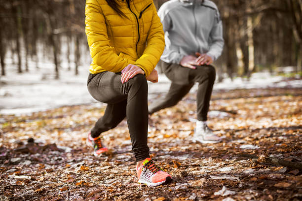 feche-se da garota de fitness forma no sportswear, fazendo a perna alongamento com seu personal trainer na floresta da manhã ensolarada de inverno. - couple stretching running jogging - fotografias e filmes do acervo