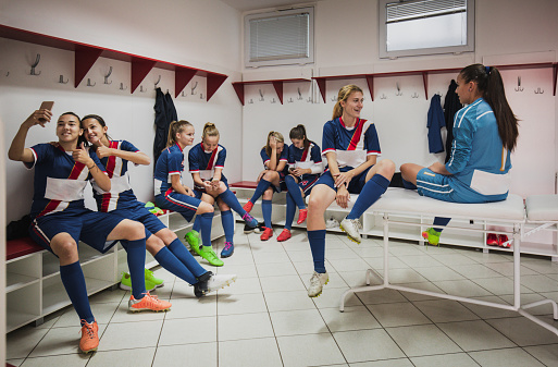 Large group of teenage soccer players relaxing in dressing room while using mobile phones and talking.