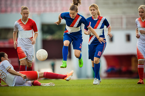 Female soccer players playing the match on a stadium while one of them is jumping over her opponent.