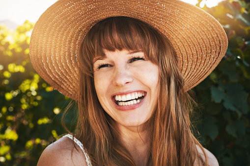 A lovely young woman with long red hair  ad very fair skin laughs happily as she stands in the sunshine wearing a sunhat.