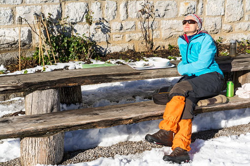Side view of senior woman while resting after worm lunch during long cross-country trekking on Mirna Gora mountain in Slovenia. On table are empty dishes, the land is covered with snow while in background is her backpack and wall of mountain cabin.