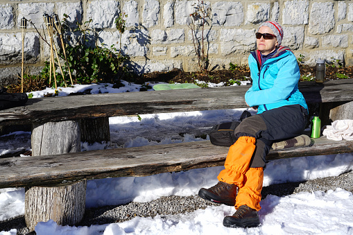 Front view of senior woman while resting after long cross-country trekking on Mirna Gora mountain in Slovenia. The land is covered with snow while in background is wall of mountain cabin.