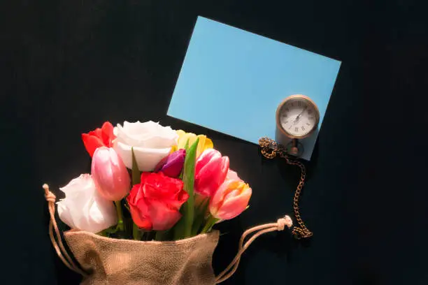 Colorful bouquet of diverse flowers in a jute bag, a vintage pocket watch and a blank blue message card, on a black background.