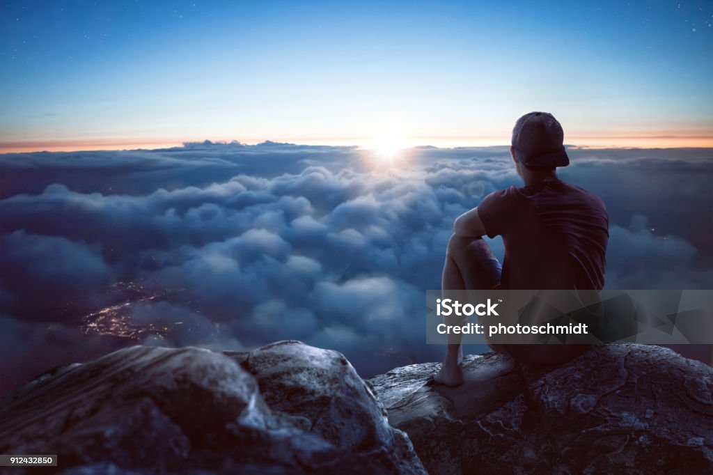 Young man with view over the clouds A young man is sitting on a rock on a summit overlooking a vast ocean of clouds. The sun is low and it is dark around him. Through the clouds you can see the lights of a city underneath. People Stock Photo