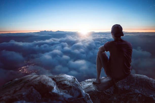 Young man with view over the clouds A young man is sitting on a rock on a summit overlooking a vast ocean of clouds. The sun is low and it is dark around him. Through the clouds you can see the lights of a city underneath. above cloud stock pictures, royalty-free photos & images