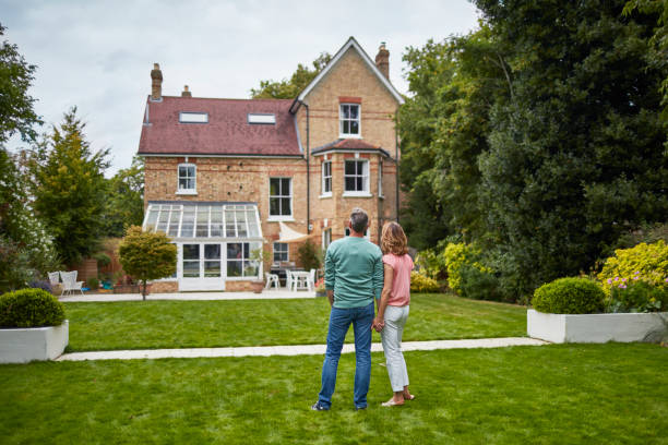 Rear view of couple on grass looking at house Rear view of mature couple looking at house. Man and woman are holding hands while standing on grassy field. They are wearing casuals. in front of stock pictures, royalty-free photos & images