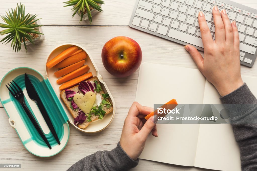 Woman eating healthy dinner from lunch box at her working table Healthy snack at office workplace. Businesswoman eating organic vegan meals from take away lunch box at wooden working table with computer keyboard, top view Snack Stock Photo