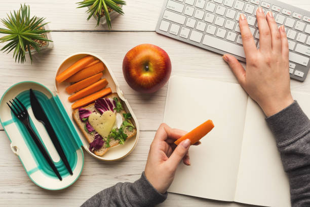 Woman eating healthy dinner from lunch box at her working table Healthy snack at office workplace. Businesswoman eating organic vegan meals from take away lunch box at wooden working table with computer keyboard, top view apple keyboard stock pictures, royalty-free photos & images