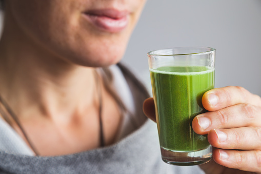 Healthy woman holding small glass of green wheatgrass juice about to take a sip.