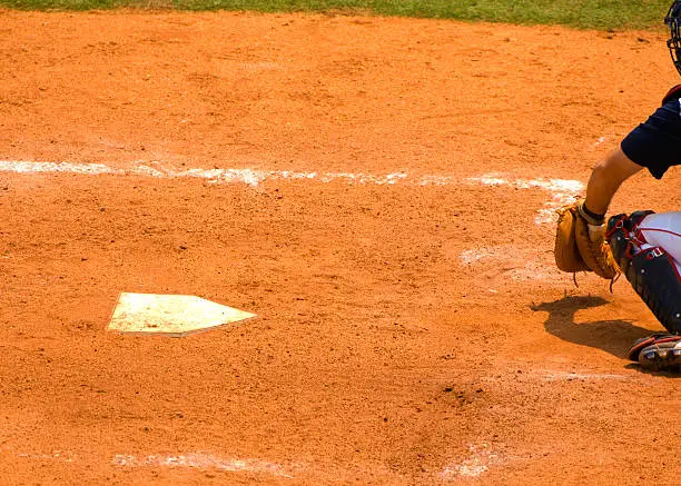 This picture is of a baseball catcher at homeplate during a baseball game catching baseballs thrown by a baseball pitcher. the catcher is squatting behind home plate. and the catcher is playing baseball in a live baseball game. the baseball player is wearing official baseball uniform and equipment and glove. the baseball player also has on safety equipment to help reduce the risk of injury. 