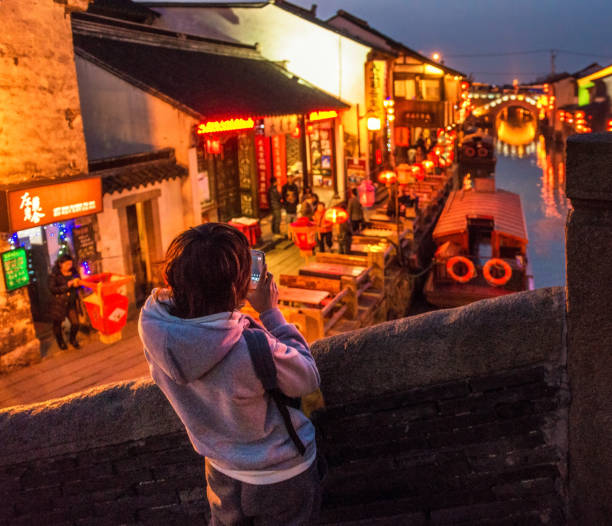 Suzhou, China - A woman on a bridge using her smartphone to photograph a traditional canal in Suzhou, illuminated at dusk by chinese lanterns.
