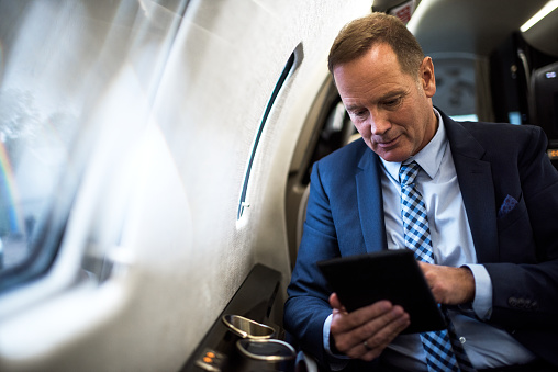 Elegant senior man sitting inside private jet airplane and holding digital tablet.