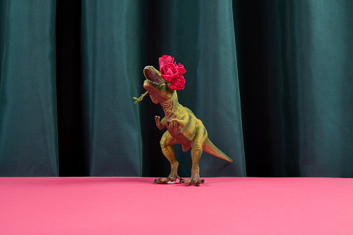a plastic toy tyrannosaurus rex eating a bouquet of red roses on a vibrant green curtain and pink background. Minimal funny and quirky pop still life photography