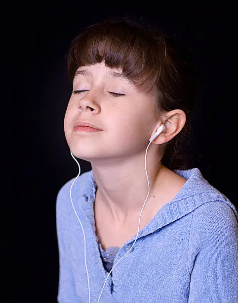 A dark haired adolescent girl listening to music with her eyes closed.  Her dark hair blends into the black background.  She is wearing white earbud style headphones.