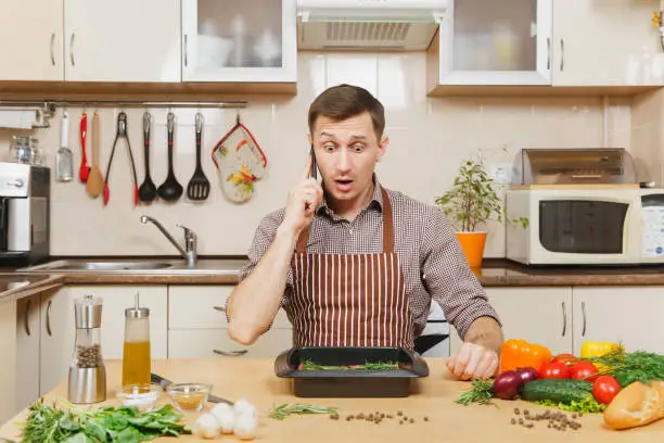 Photo of Perturbed stress young man in apron sitting at table with vegetables, talking on mobile phone, cooking at home preparing meat stake from pork, beef or lamb, in light kitchen with wooden surface.