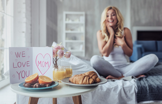 We love you, mom! Attractive young woman is sitting on bed at home. Breakfast in bed on Mother's Day with a children postcard.