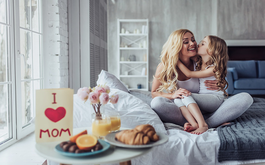 Attractive young woman with little cute girl are spending time together at home while sitting on bed. Happy family concept. Breakfast in bed on Mother's day.