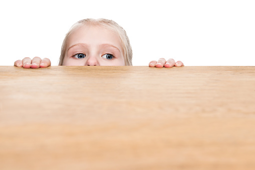 Child sticks his eyes out from under the table and looks sideways, isolated on a white background