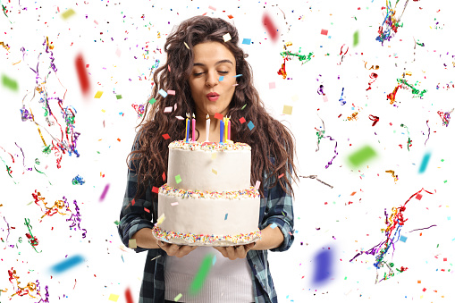 Teenage girl blowing candles on a birthday cake with confetti streamers flying around her isolated on white background
