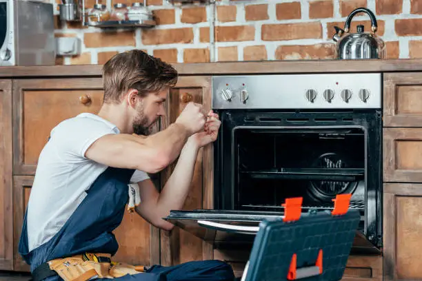Photo of young repairman in protective workwear fixing oven in kitchen