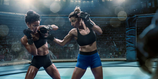 Professional Female Mixed Martial Arts Fighters Throw Punches in Octagon Two professional female mixed martial arts fighters competing in an octagon inside an indoor floodlit arena. Both fighters are in side on stance, and dressed in tight shorts and sports bras. One fighter has attacked with a jab or uppercut which has been blocked by her opponent. fighting stance stock pictures, royalty-free photos & images
