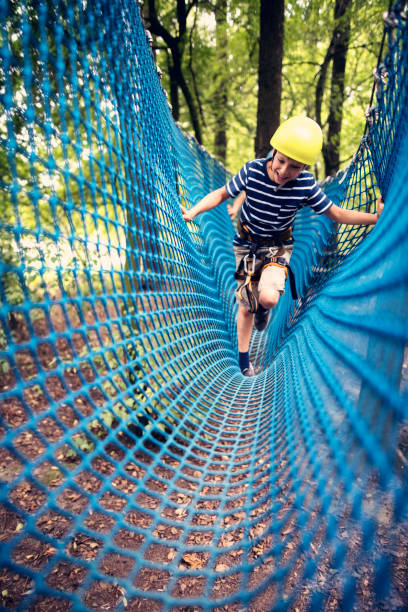 Little boy wearing a helmet walking through difficult obstacle in ropes course in adventure park. The laughing boy is crossing a net.