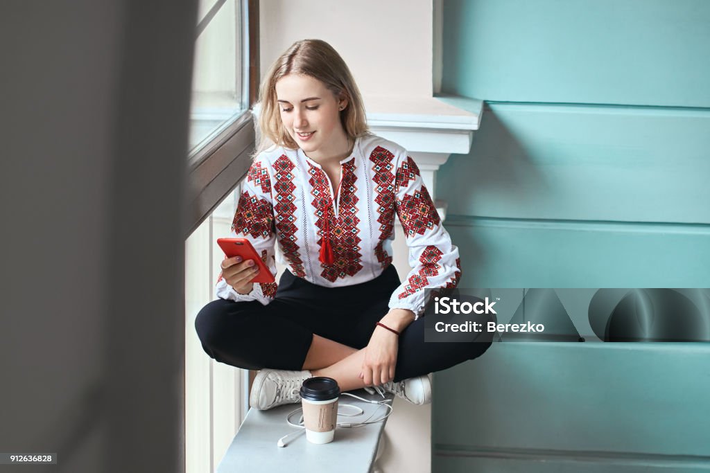 Young beautiful caucasian girl student wearing a vyshyvanka, a traditional Ukrainian embroidered shirt checks her smartphone at university Young beautiful caucasian girl student wearing a vyshyvanka, a traditional Ukrainian embroidered shirt checks her smartphone while drinking coffee between lectures Ukrainian Culture Stock Photo