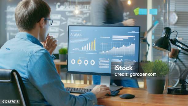 Analyst Works On A Personal Computer Showing Statistics Graphs And Charts In The Background His Coworker And Creative Office Stock Photo - Download Image Now